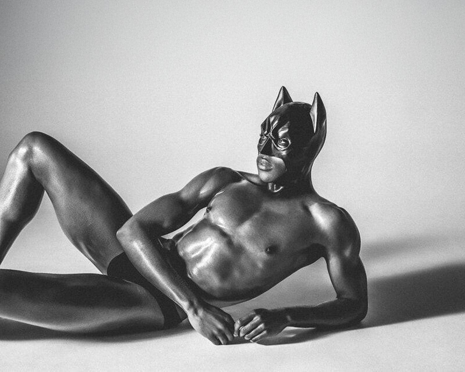 A muscular figure in a sleek black mask reclines against a light background, showcasing a bold and dramatic pose.
