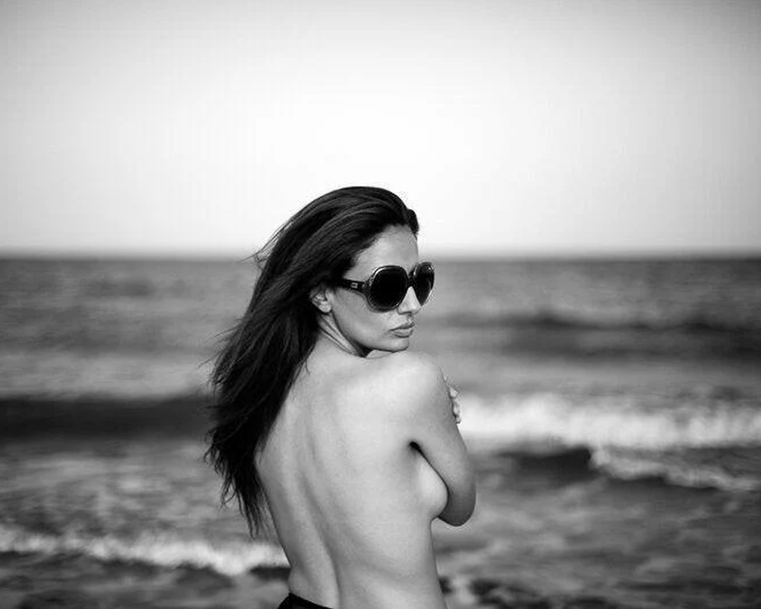 Black and white photo of a woman with sunglasses standing on a beach.