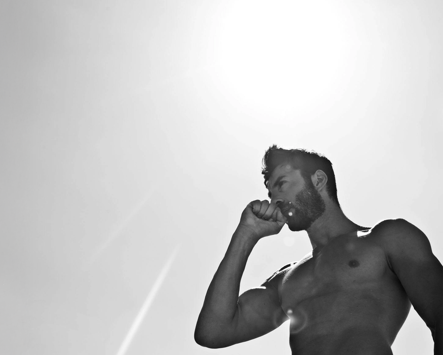 A muscular man stands against a bright sky, sipping from a drink with the sun shining overhead in a black and white setting.