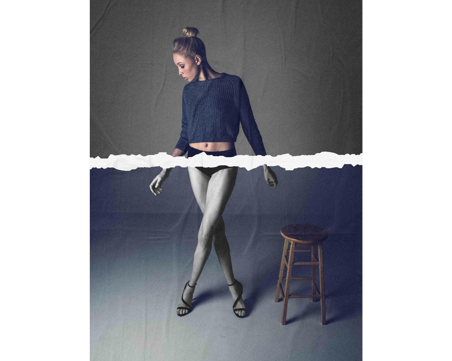 A model stands gracefully, wearing a cropped sweater and shorts, with one leg forward, beside a wooden stool against a textured backdrop.