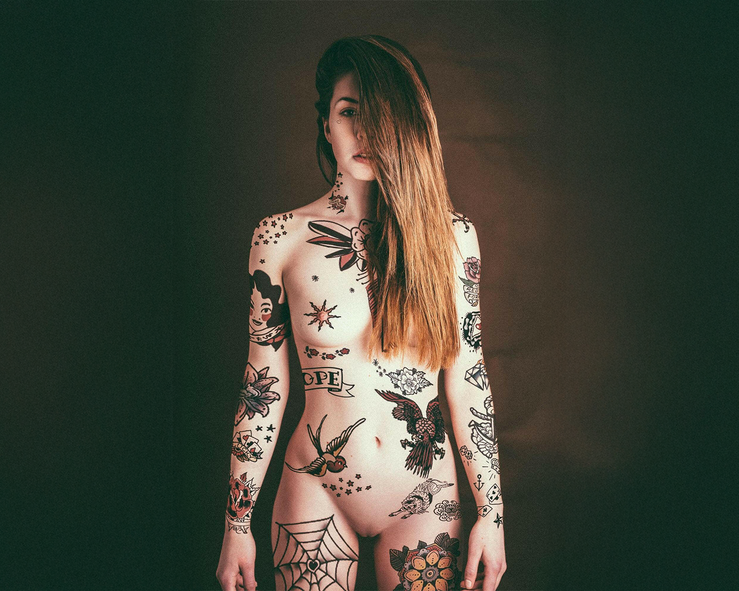 A woman with intricate tattoos covering their body, featuring designs like flowers, stars, and vintage motifs, against a dark background.