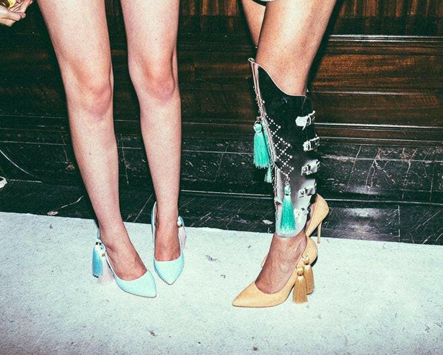 Two pairs of stylish high-heeled shoes: one in light blue with tassels, one yellow with bold embellishments, alongside bare legs.