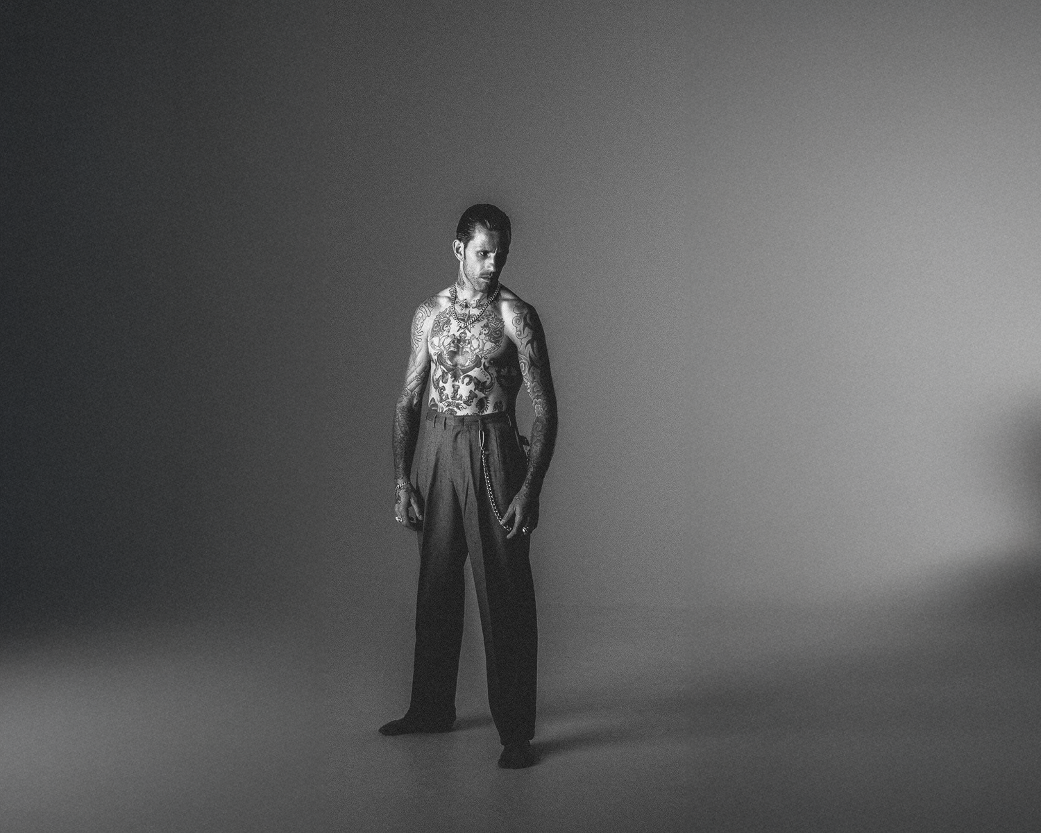 A tattooed man is positioned in a shadowy room, wearing a pant, with the darkness adding to the scene's intrigue