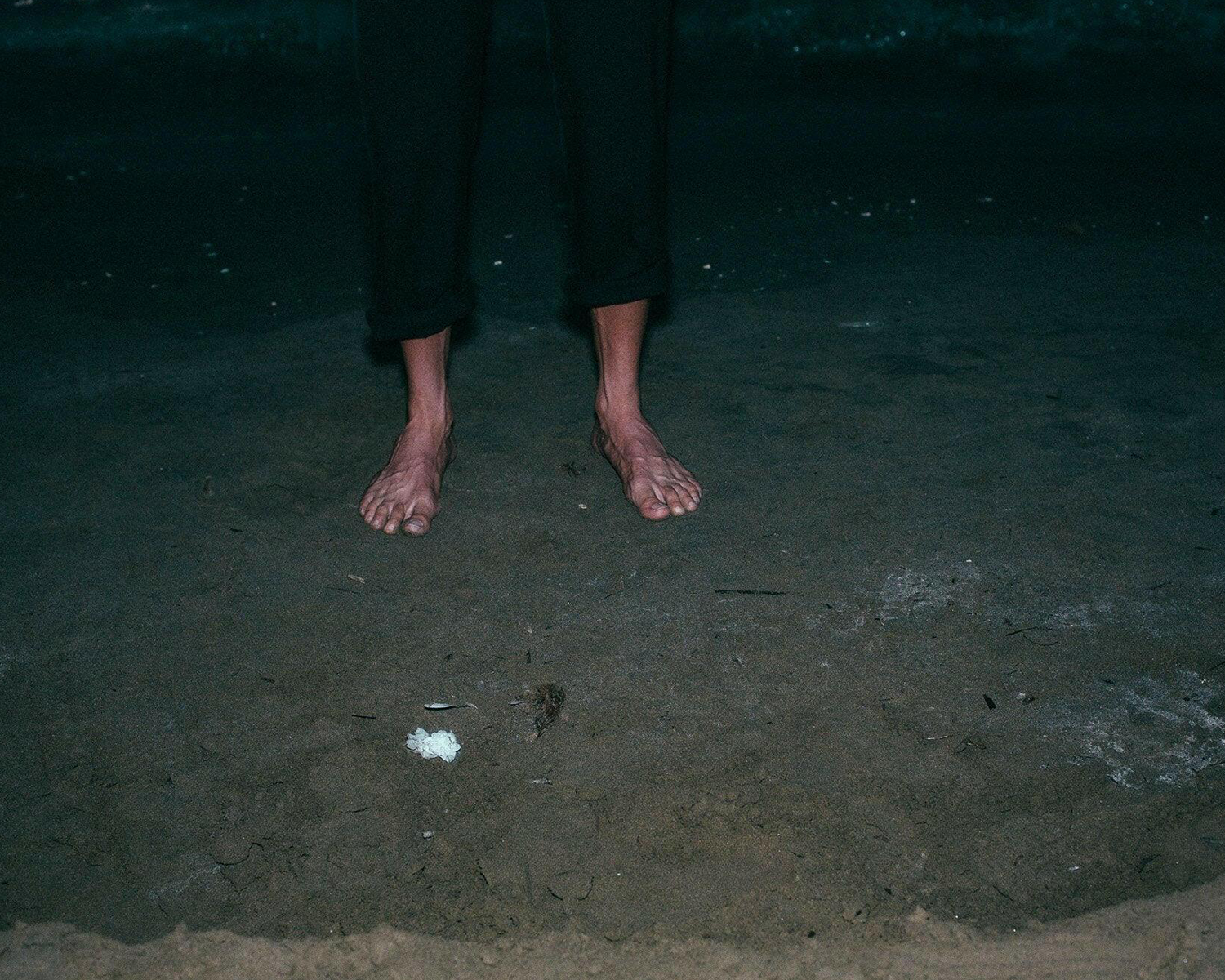 Feet of a person standing on a dark sandy surface with minimal lighting.