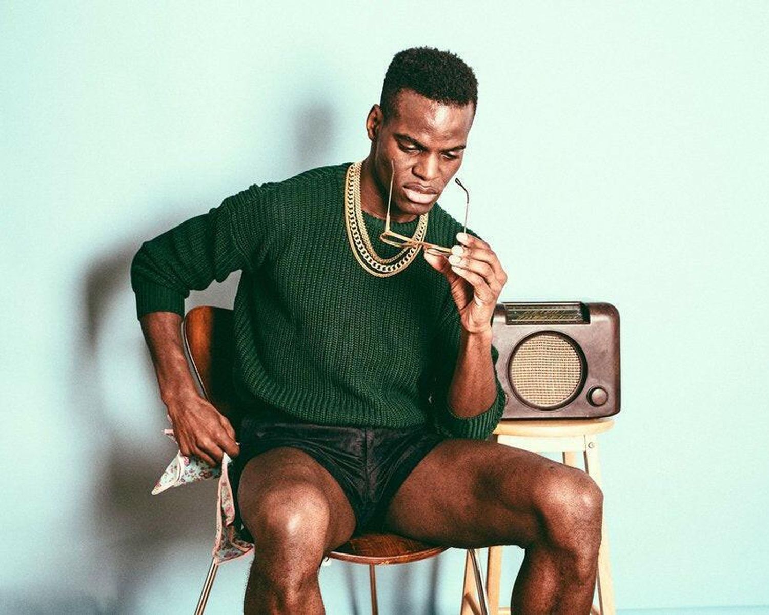 Man sitting on a stool with a vintage radio, wearing a green sweater and black shorts.