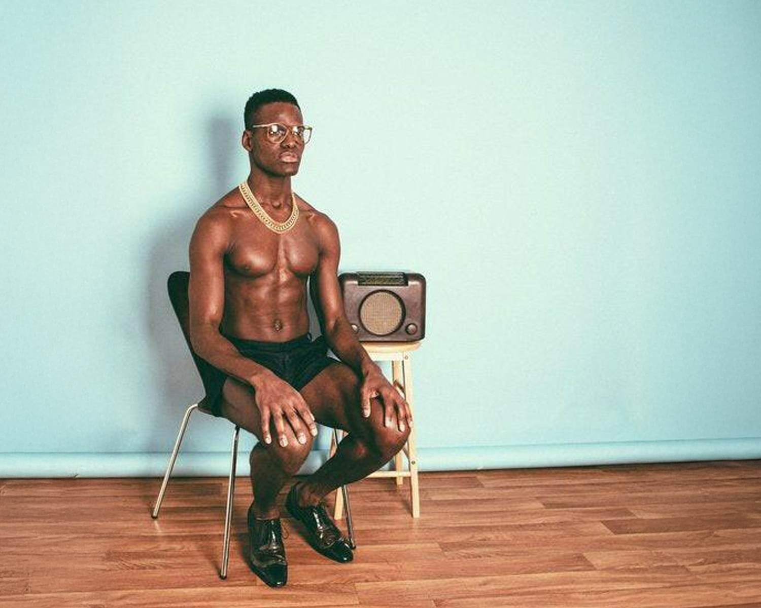 Man sitting shirtless on a chair with a radio in a room with light blue walls and wooden floor.