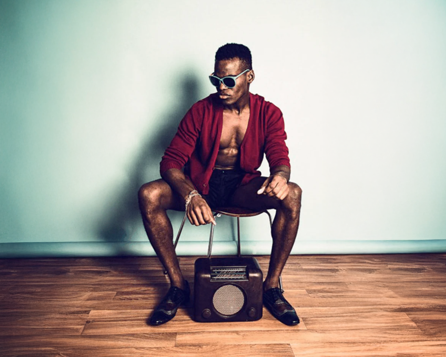 Man in red jacket sitting on a stool with a vintage radio, against a plain wall.
