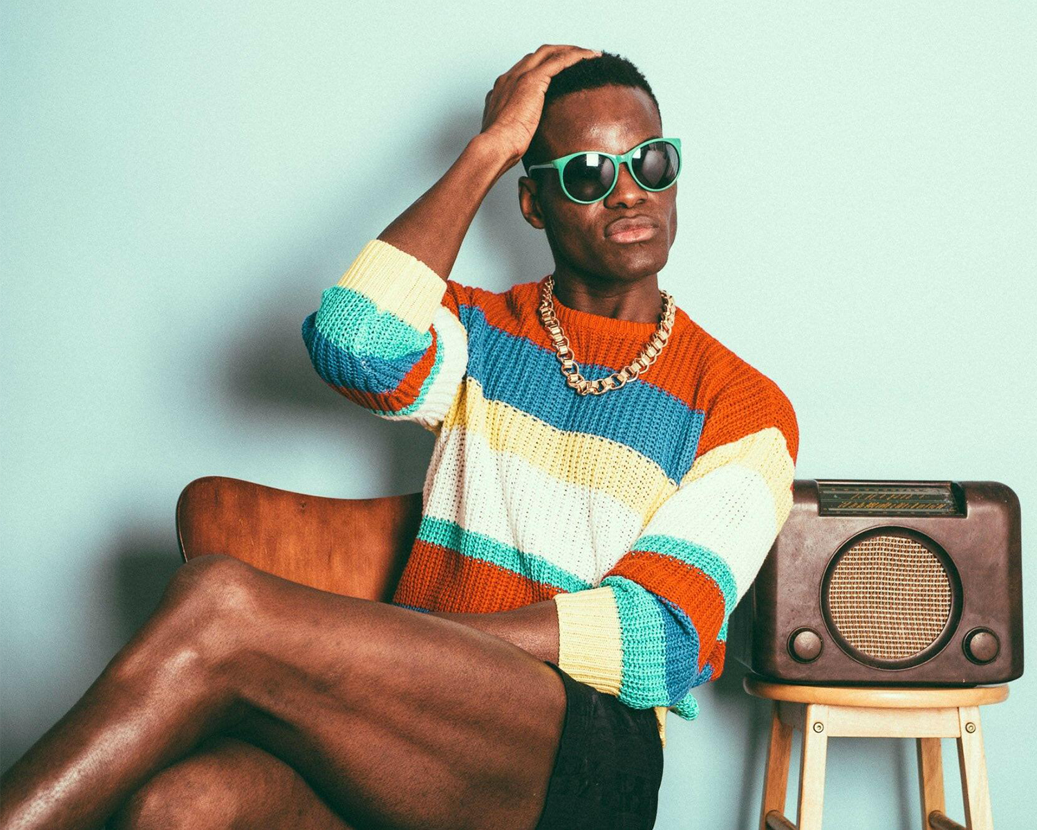 Man wearing a colorful striped sweater and sunglasses sitting on a chair with a radio.