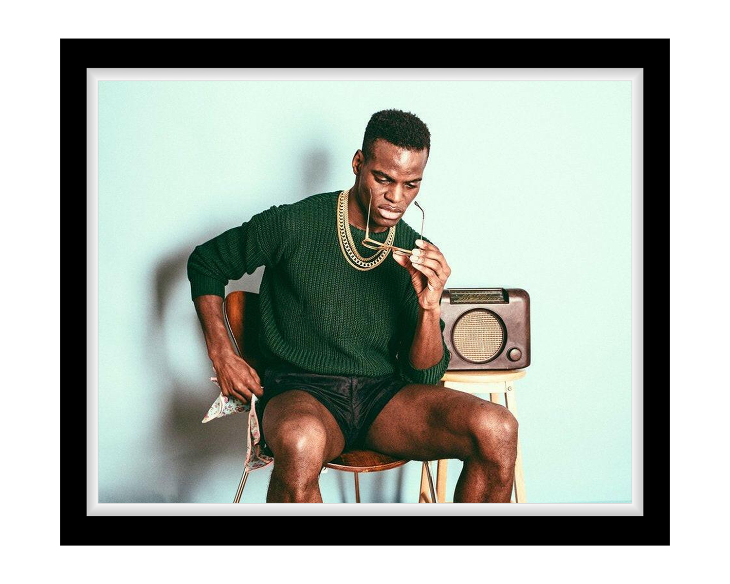 Man sitting on a stool with a vintage radio and newspaper, wearing a green sweater and gold chains.