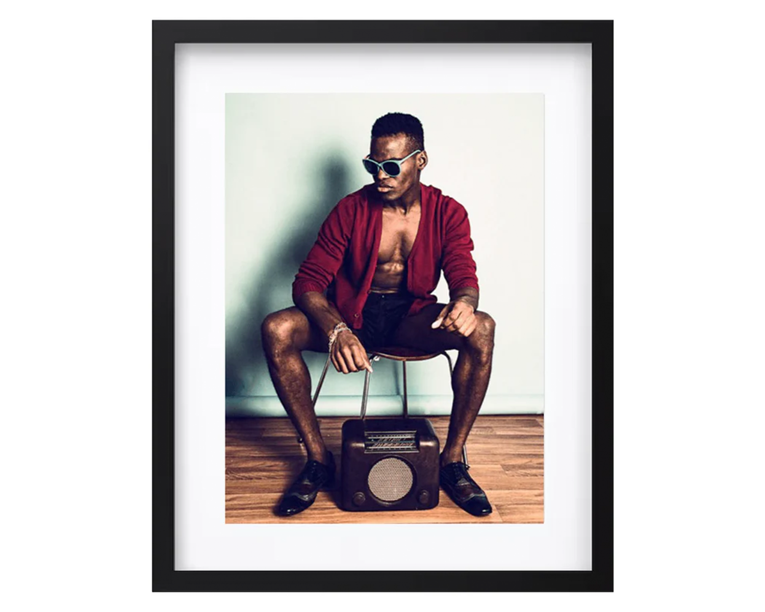 Man sitting on a stool with a boombox, wearing sunglasses and a red jacket, framed in black.