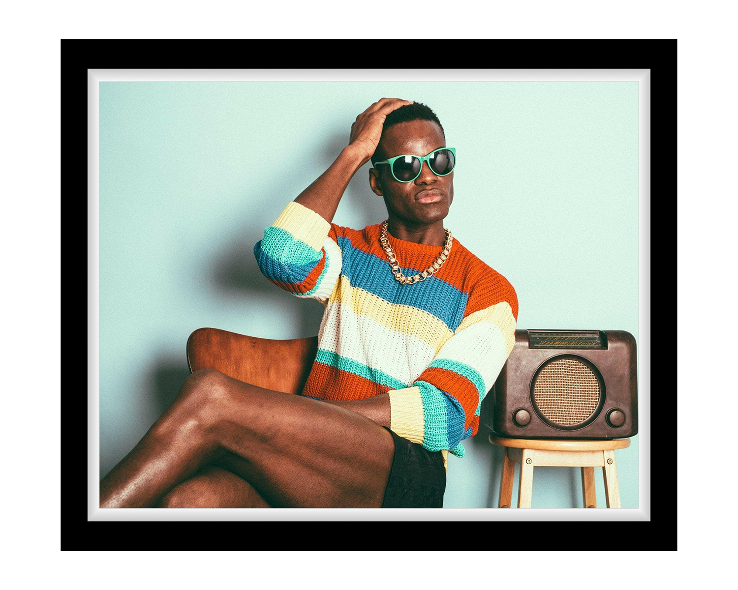 Man wearing a colorful striped sweater sitting on a stool with a radio, against a light blue background.