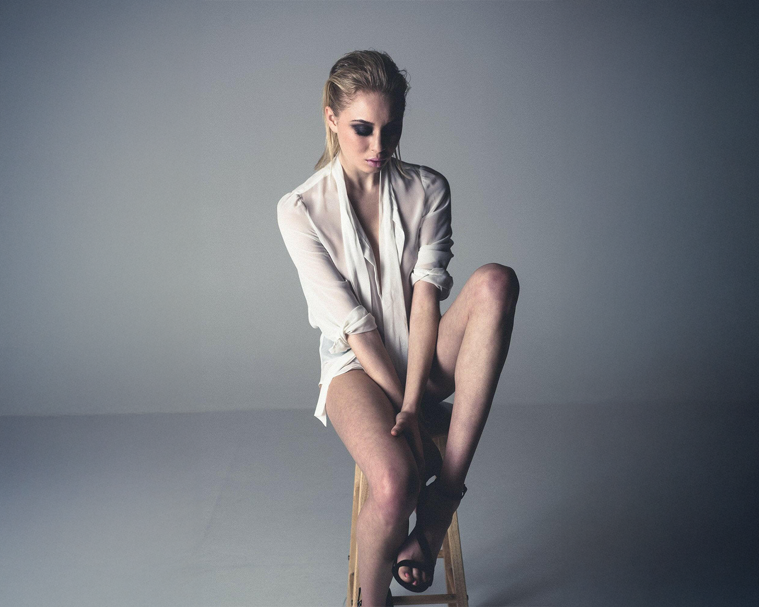 A woman in a white shirt and black shoes is seated on a stool, with a relaxed posture and a neutral expression.